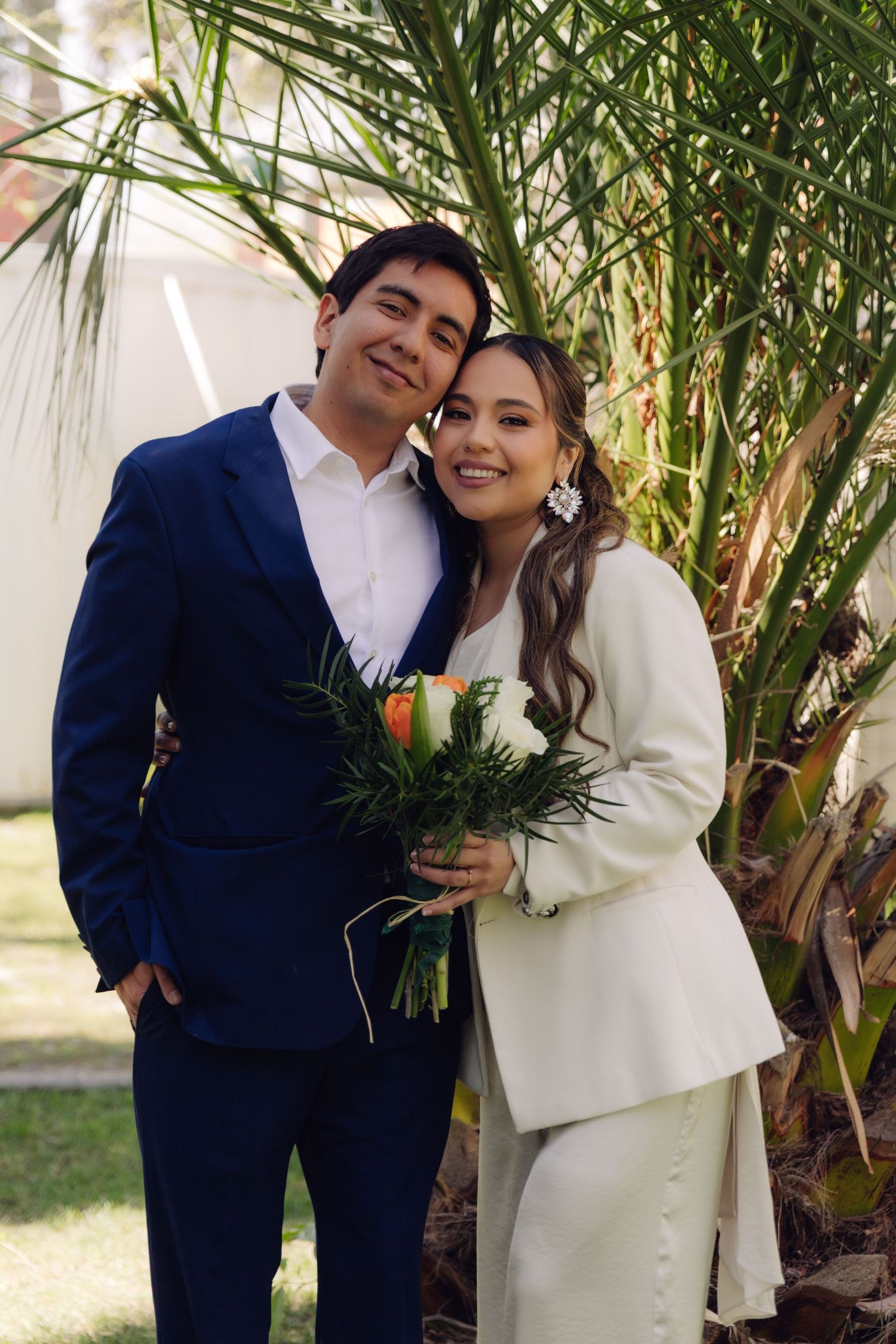 Smiling couple posing outdoors, groom in blue suit and bride in white pantsuit holding bouquet.