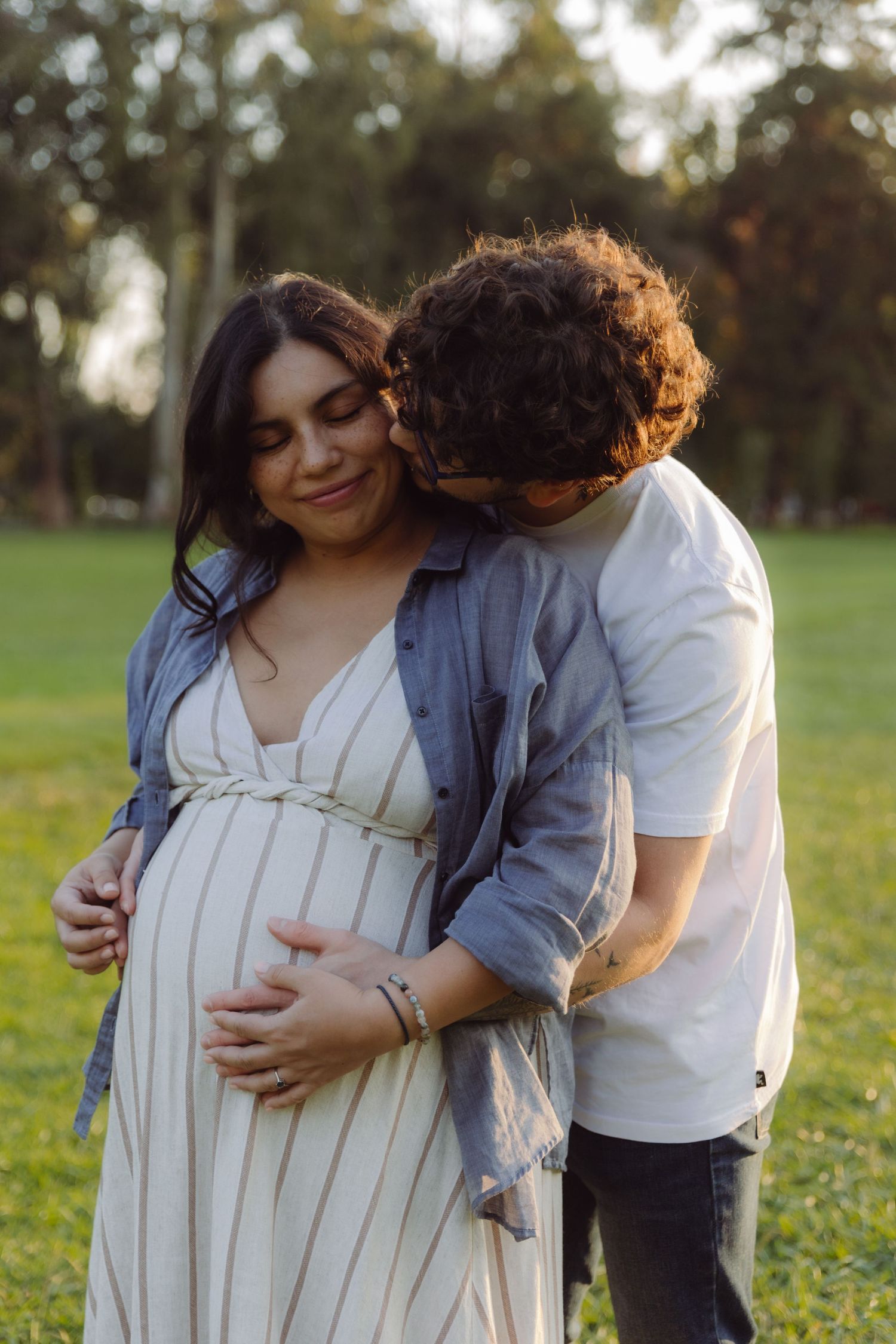 Couple sharing tender moment outdoors, man embracing pregnant woman's belly in sunlit park.