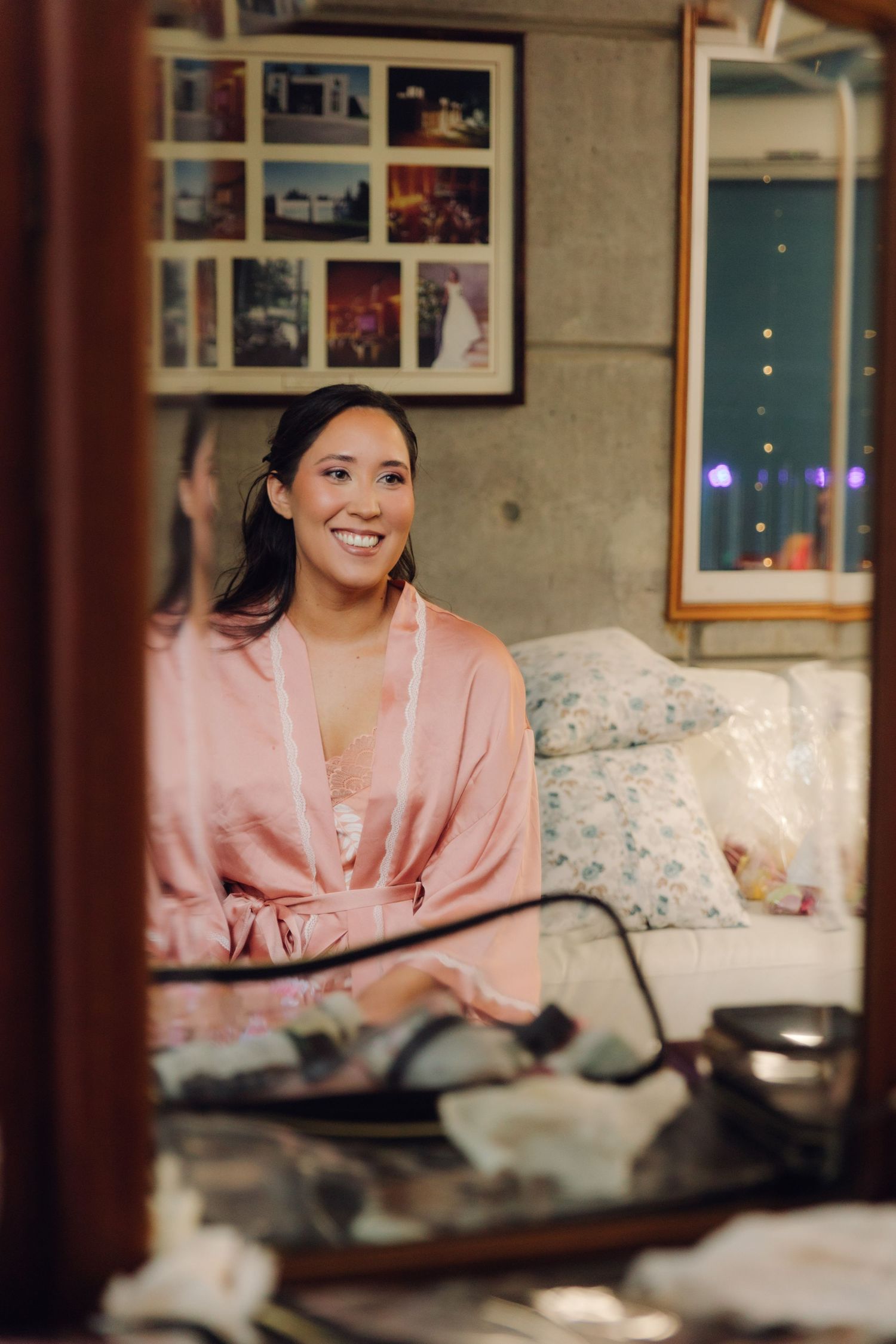 Happy bride in pink satin robe smiling at her reflection in a mirror while getting ready.