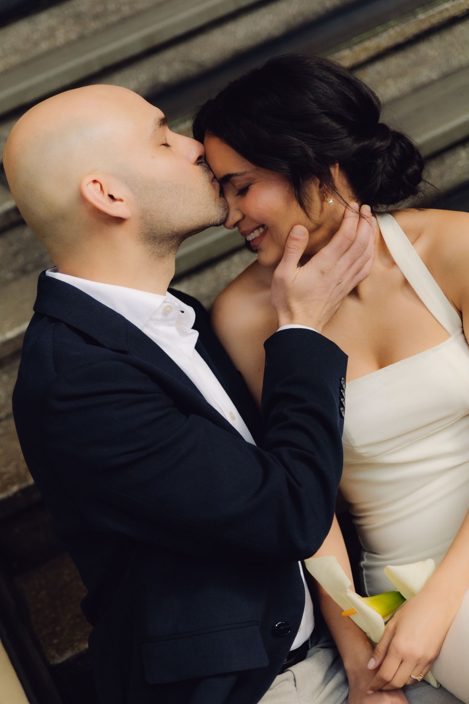 Bald groom kissing smiling bride on cheek while sitting on steps.