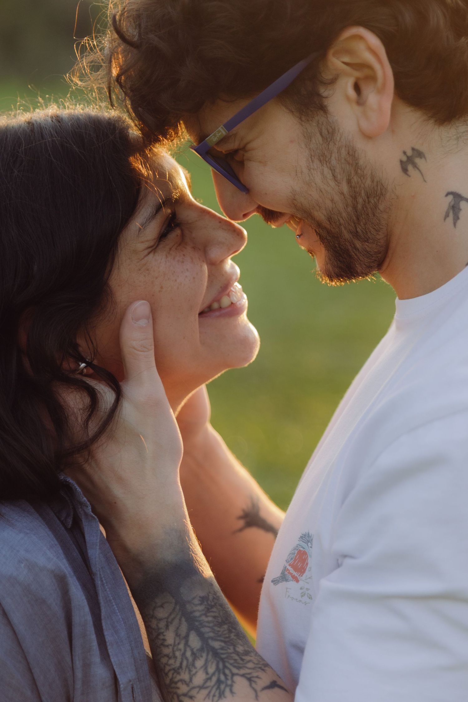 Close-up of tattooed couple almost kissing, golden hour light, man wearing blue glasses.