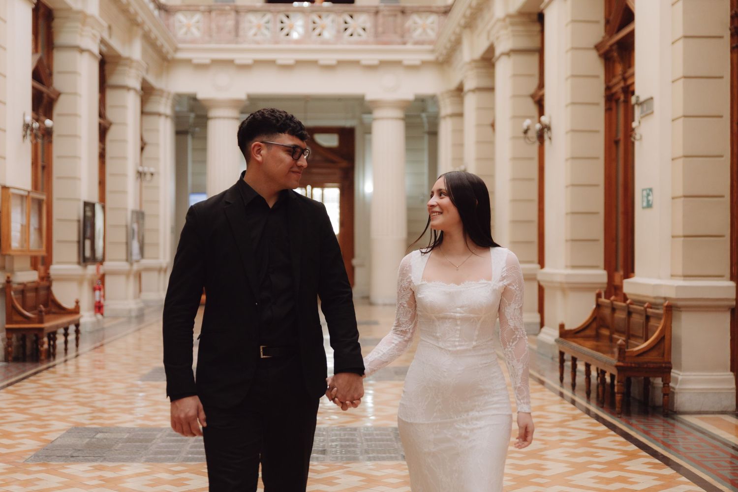 Engaged couple walks hand in hand through elegant columned hall, woman in white lace dress.