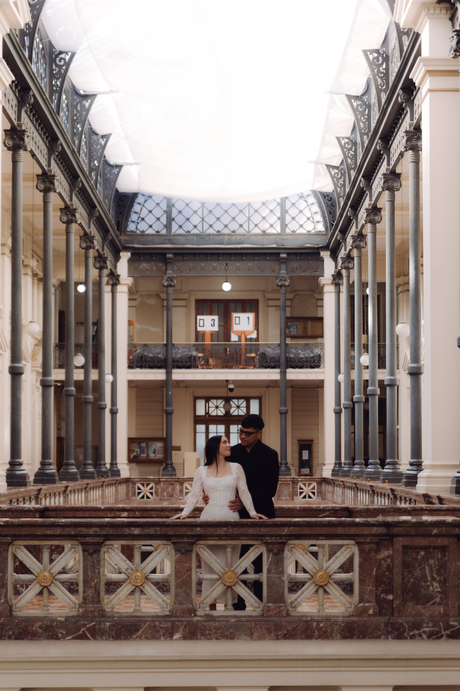 Couple posing on an ornate marble balcony inside a grand historic building with decorative iron columns.