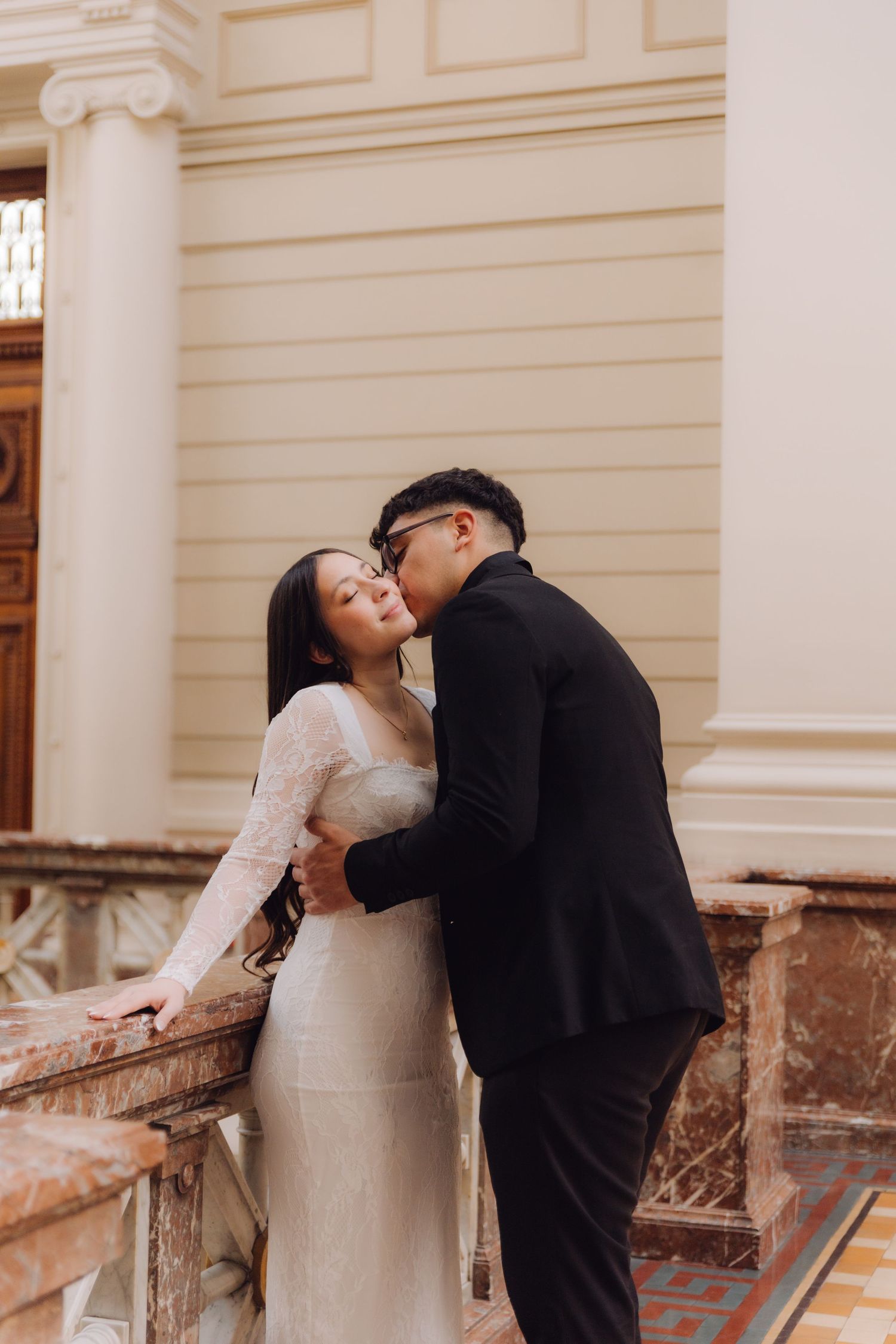 Asian couple sharing a kiss on a marble balcony inside an elegant classical architecture building.