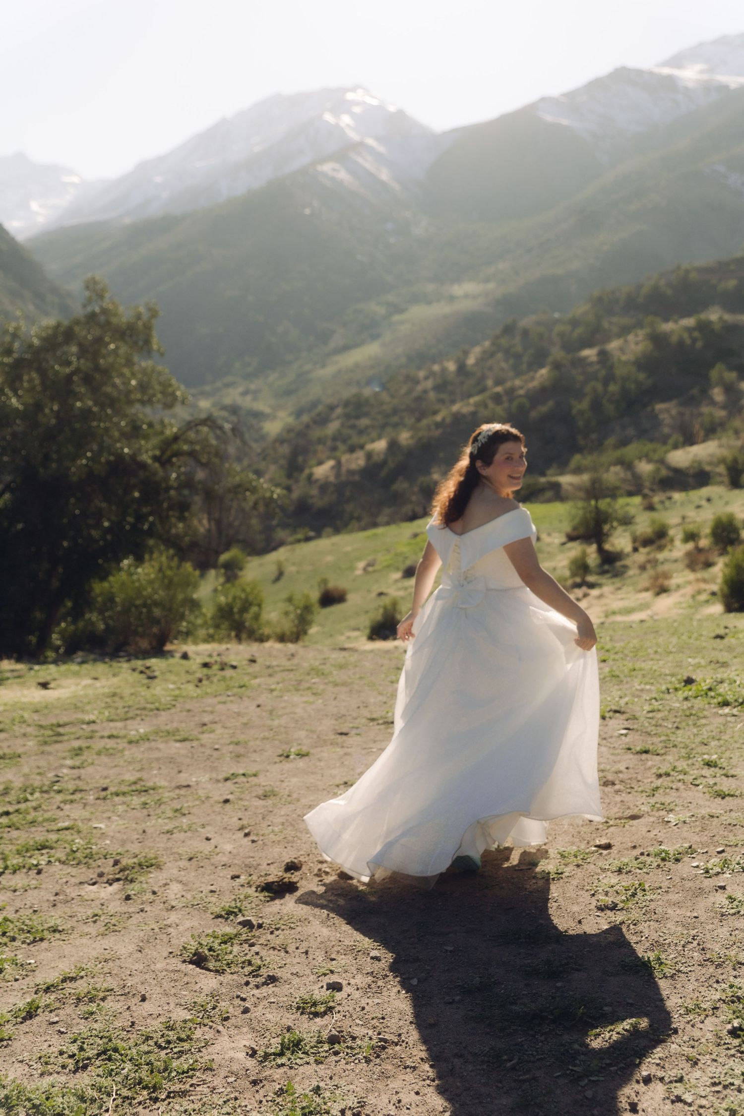 Bride in white off-shoulder wedding gown twirling gracefully in a lush green mountain meadow.