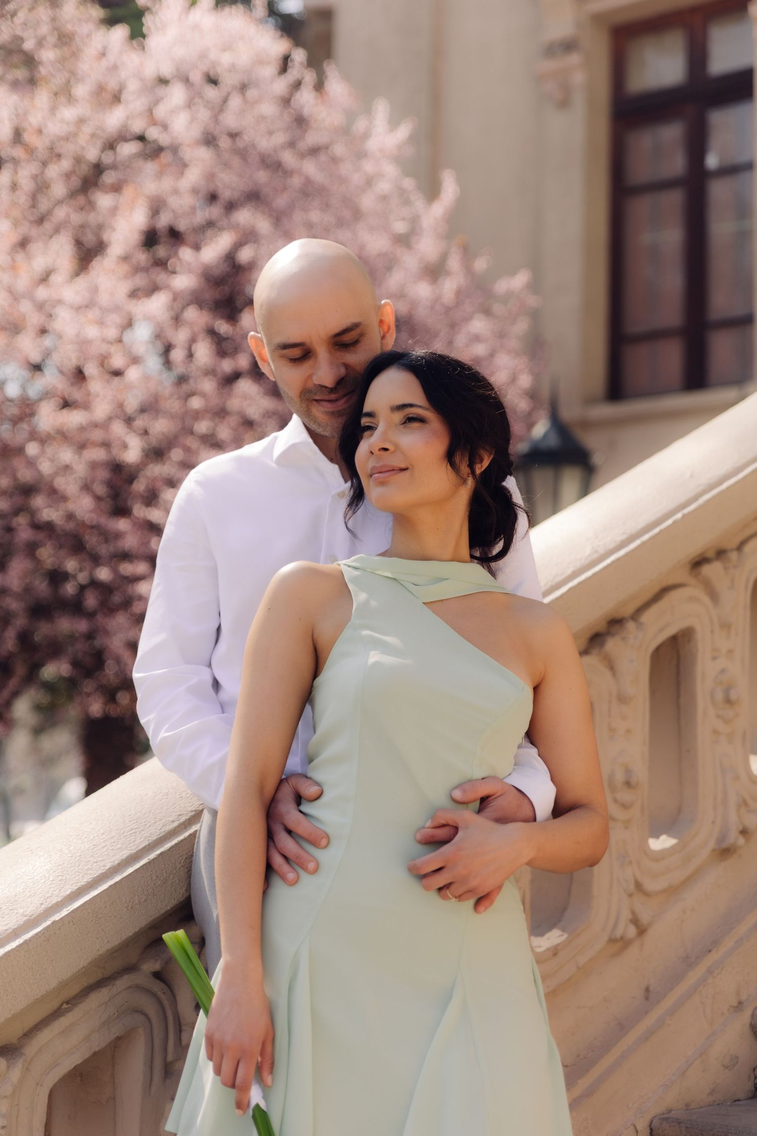 Bald man in white shirt embracing woman in mint dress on balcony.