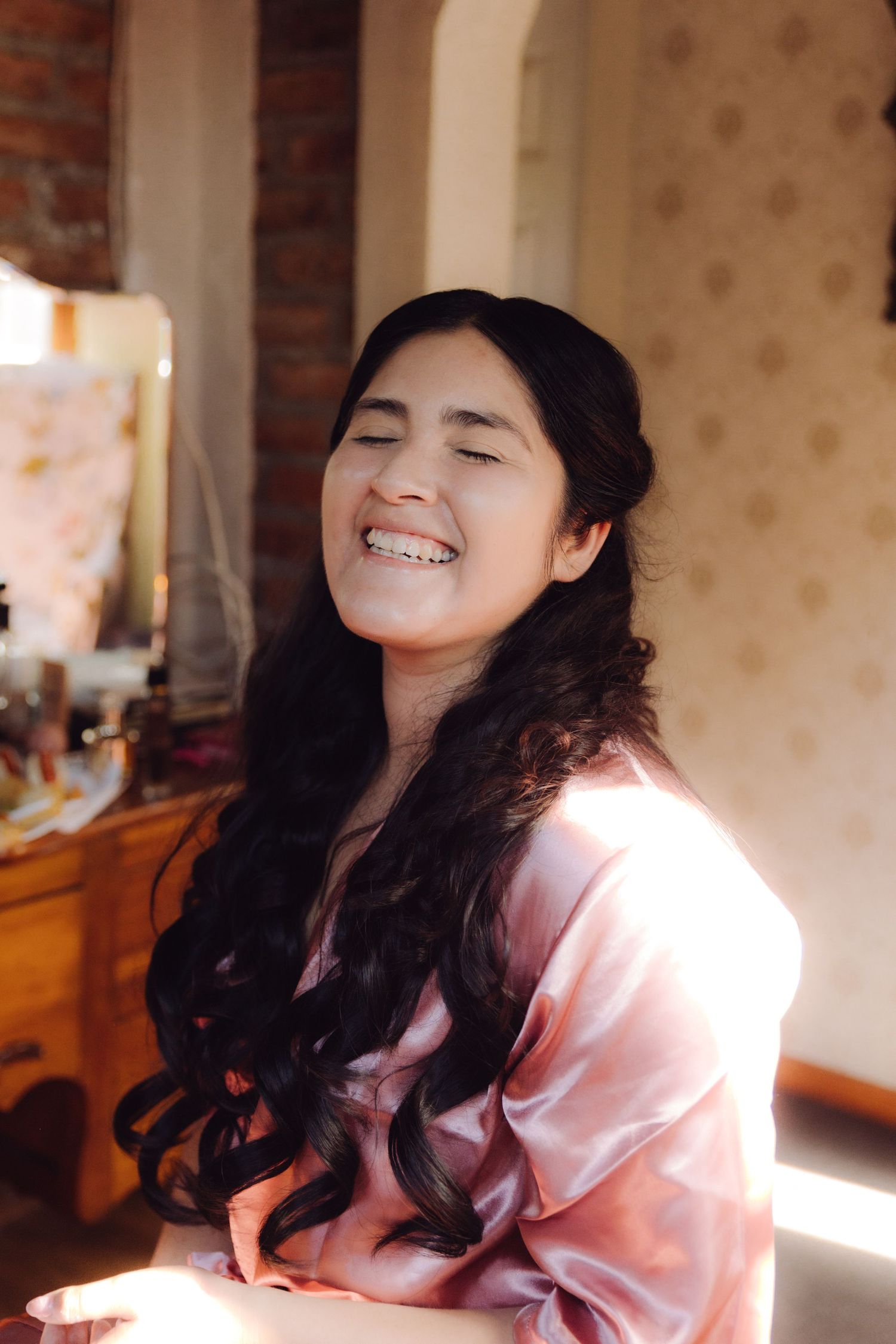 Young woman with long dark curly hair laughs joyfully in sunlight wearing pink satin robe during bridal preparations.