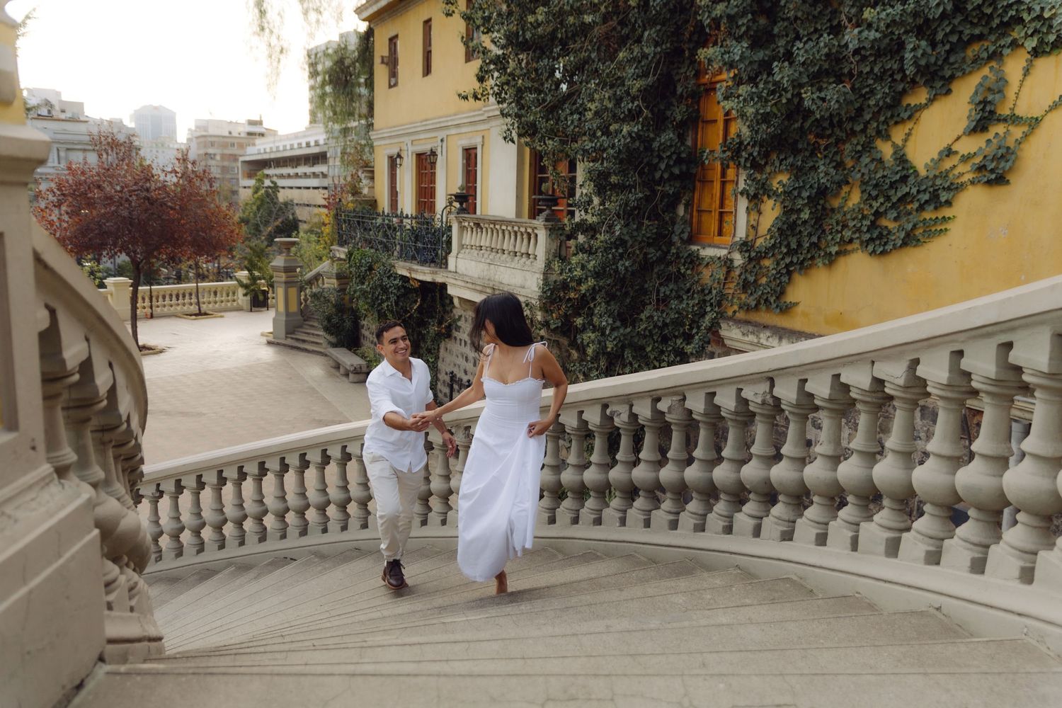 Couple in white outfits walking up ornate stone stairs beside an ivy-covered yellow building.