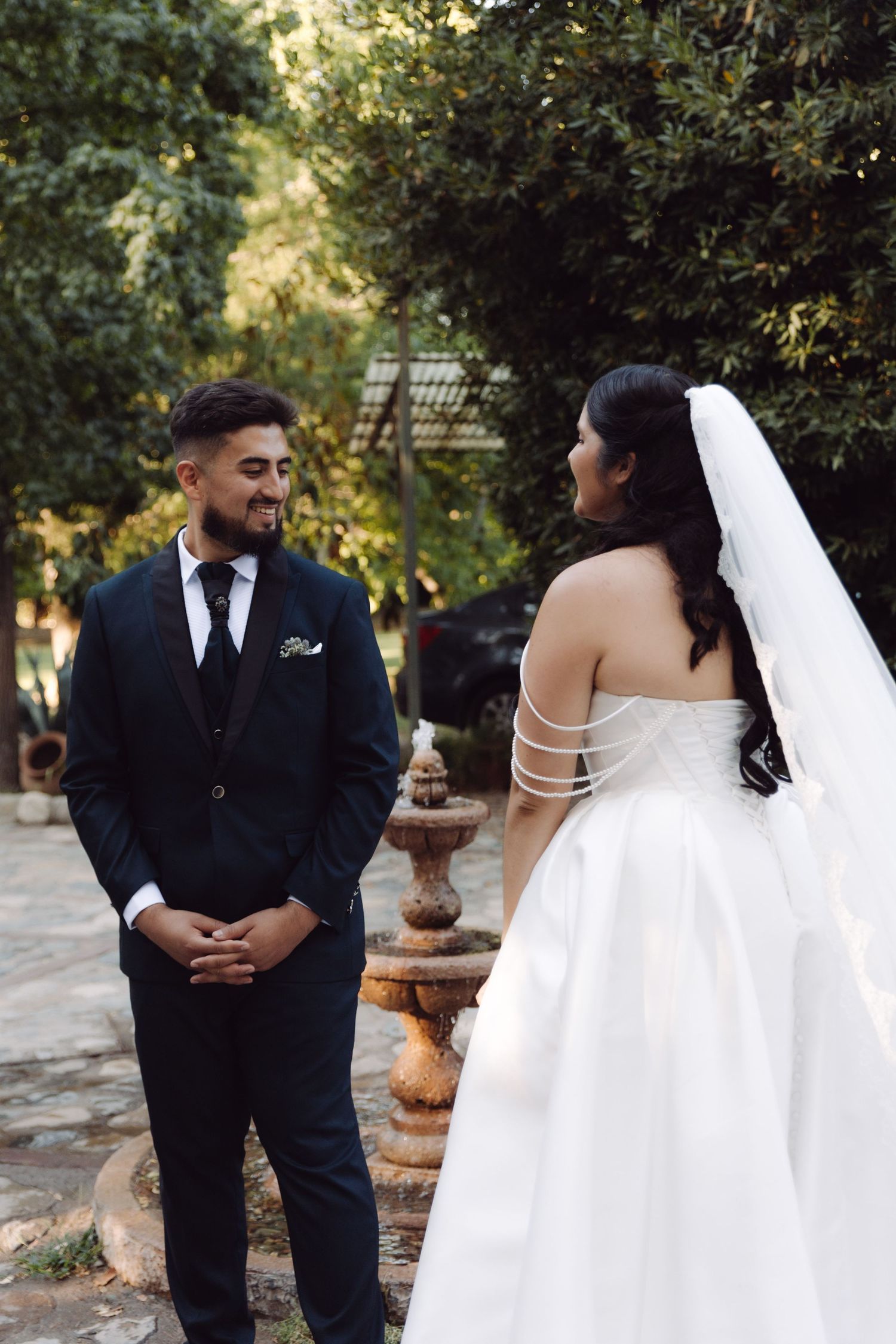 Groom in black suit smiles as he sees bride in strapless white gown with strappy back detail during first look outdoors.