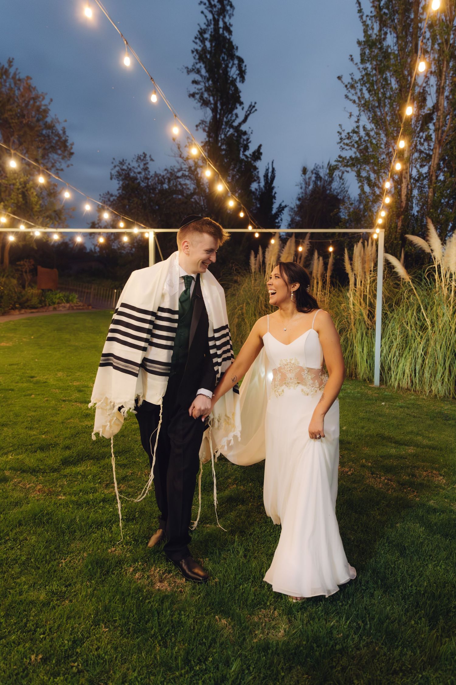 Jewish bride and groom walking hand in hand under string lights at dusk in a garden.