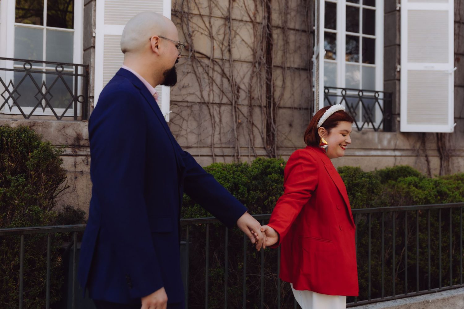 Couple holding hands while walking past a charming European building, woman in red blazer.