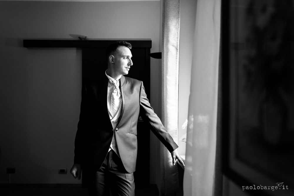Black and white portrait of a groom standing by a window in elegant attire.