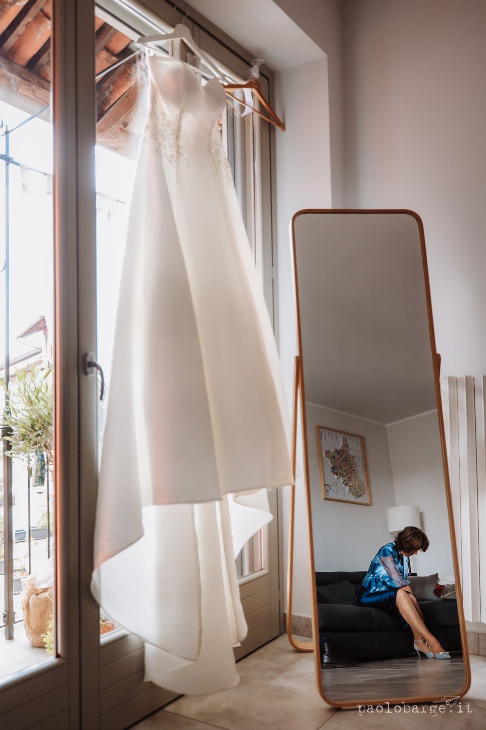 An elegant white wedding dress hangs by a window with a full-length mirror reflection.