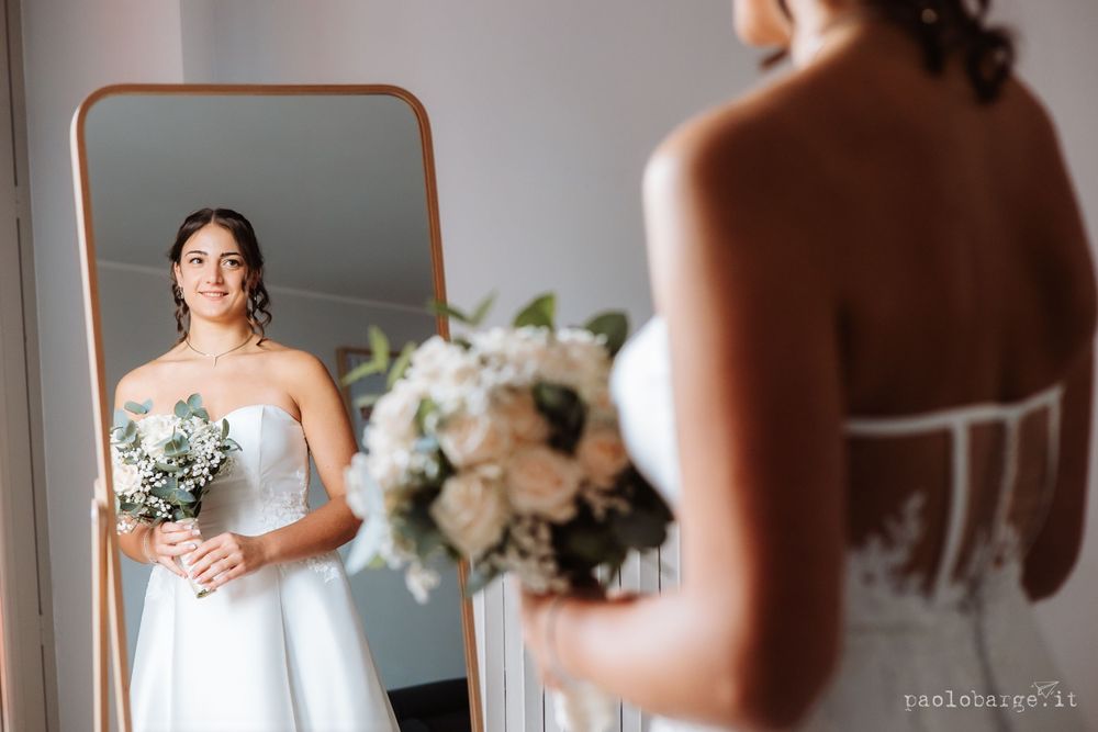A reflection in a mirror shows a person in a wedding dress holding a white floral bouquet.