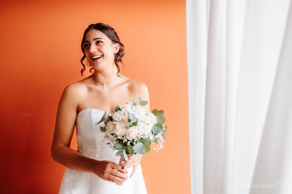 A radiant bride in white holds a delicate bouquet against an orange wall.