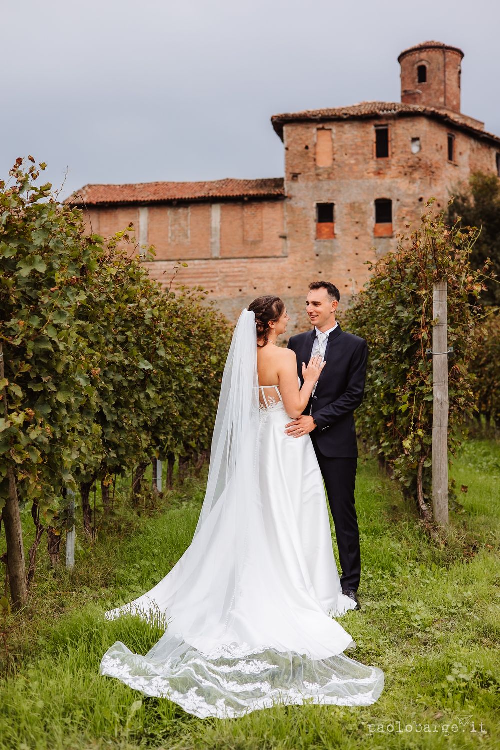 Elegant wedding couple poses in front of rustic Italian villa surrounded by vineyard.