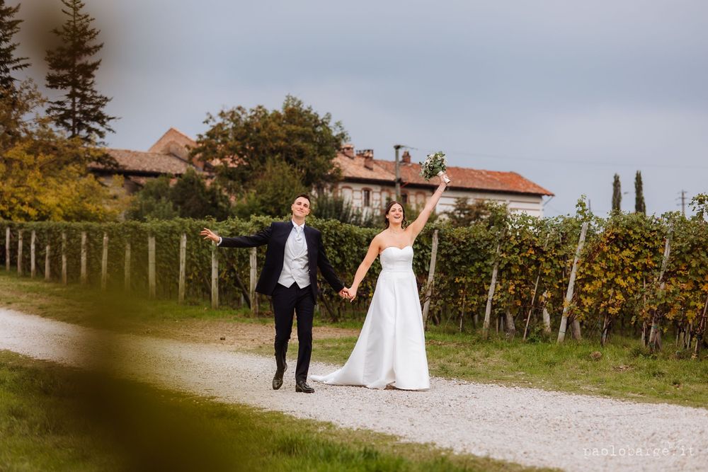 A wedding couple walks along a gravel path between vineyard rows with rustic buildings in the background.