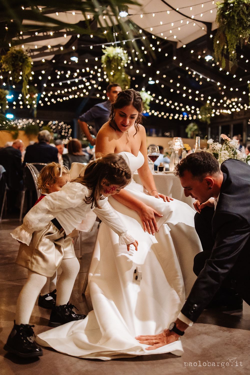 Children playfully interact with a bride's wedding dress during reception festivities.