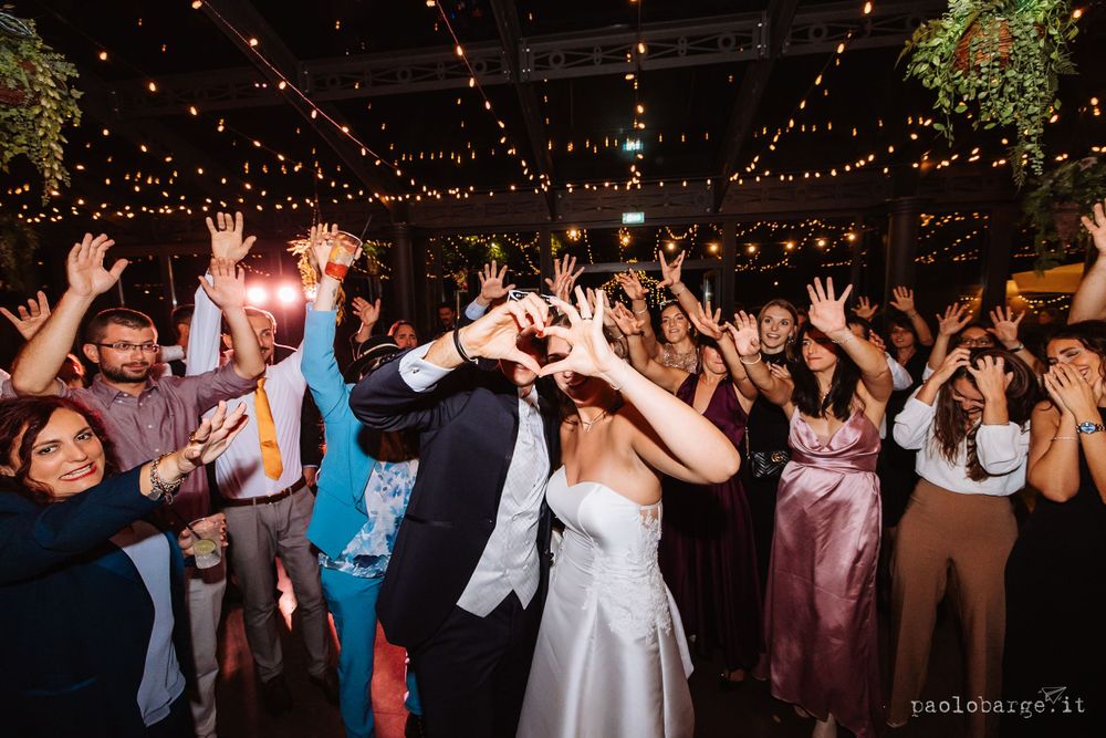 Wedding guests dance with hands in the air under twinkling string lights during an indoor reception celebration.