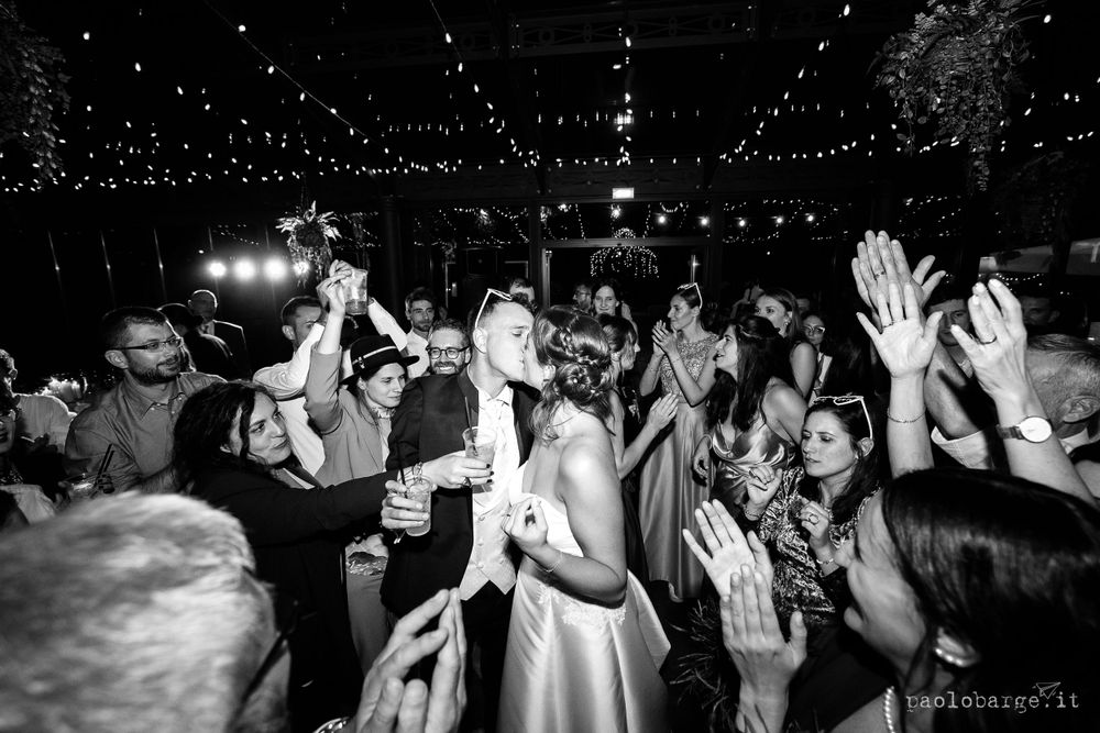 Black and white photo of a lively wedding reception dance floor with people dancing and raising hands in celebration.