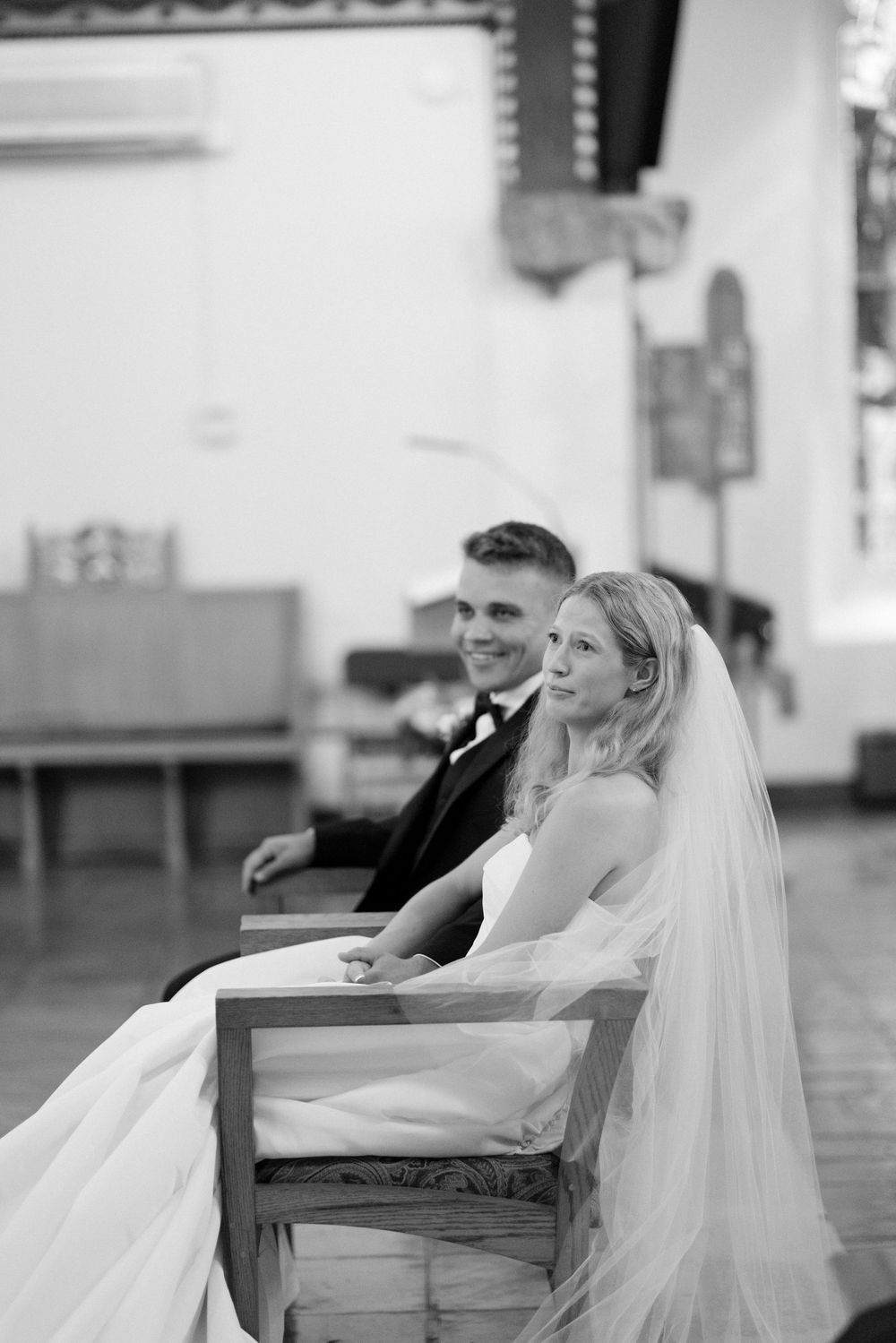 A pair in wedding attire sits together on a wooden bench in a traditional indoor setting.