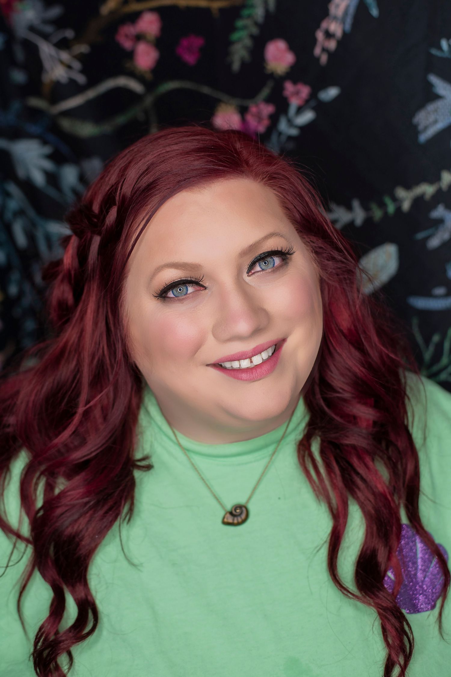Portrait of someone wearing a mint green sweater against a dark floral background, with vibrant red hair and bright eyes.