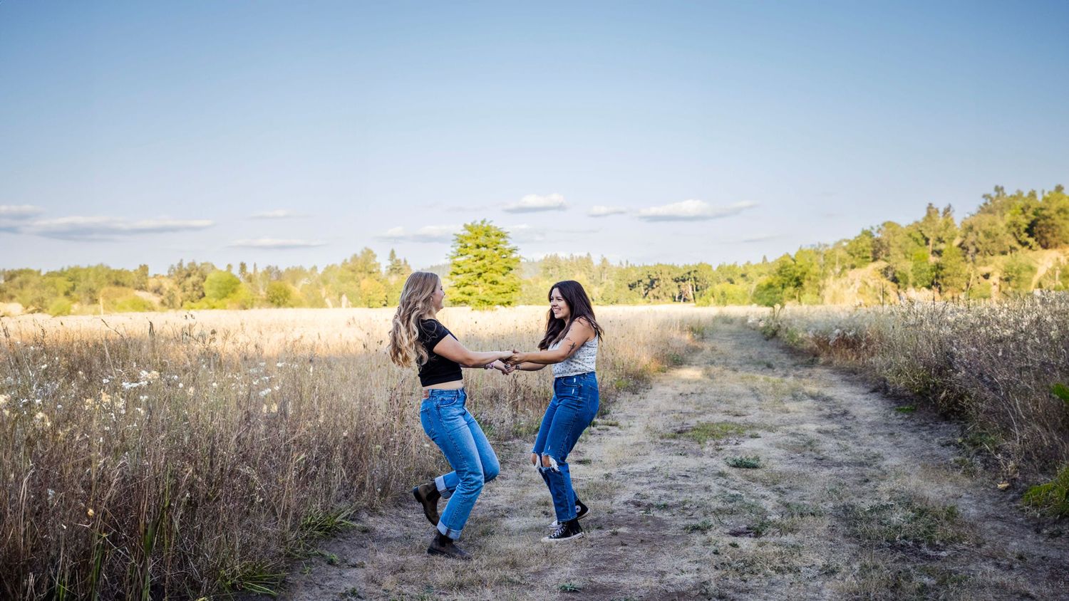 two girls hold hands and spin along a trail in a field that is golden with green trees in distance