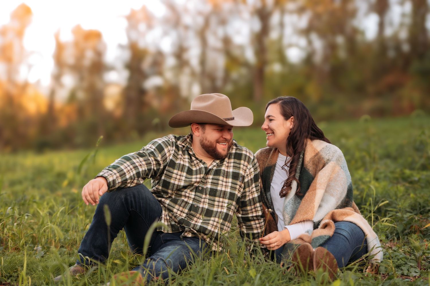 Two people wearing casual western attire share a candid moment while sitting in a grassy field at sunset.