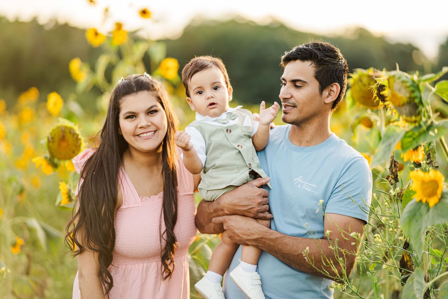 A family poses together in a sunflower field at golden hour wearing coordinating pink and blue outfits.