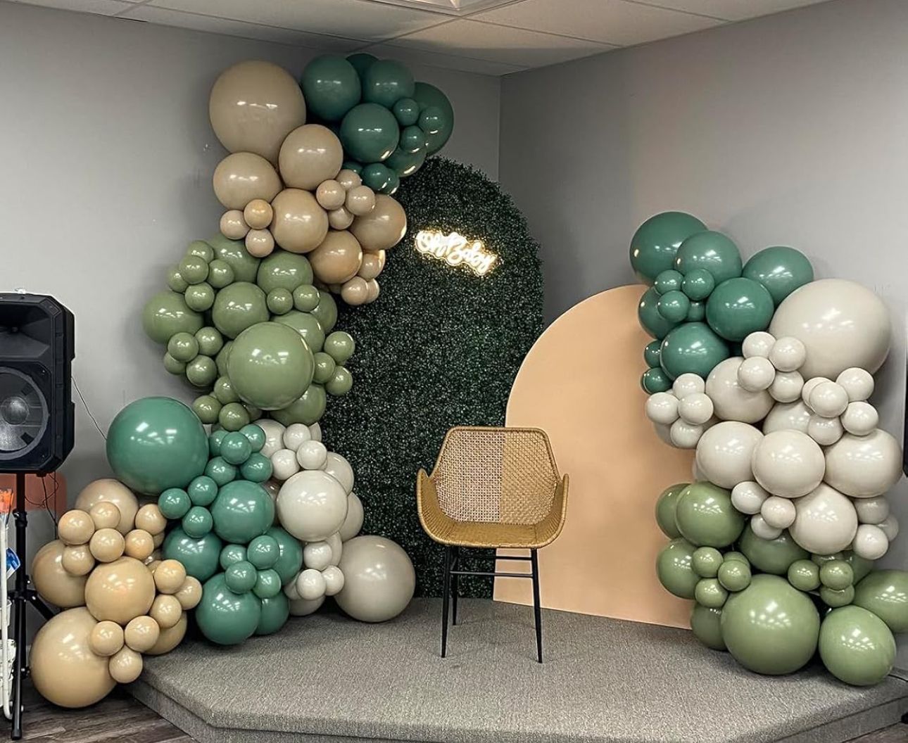 Green and beige balloon garland arch with boxwood wall backdrop and rattan chair.