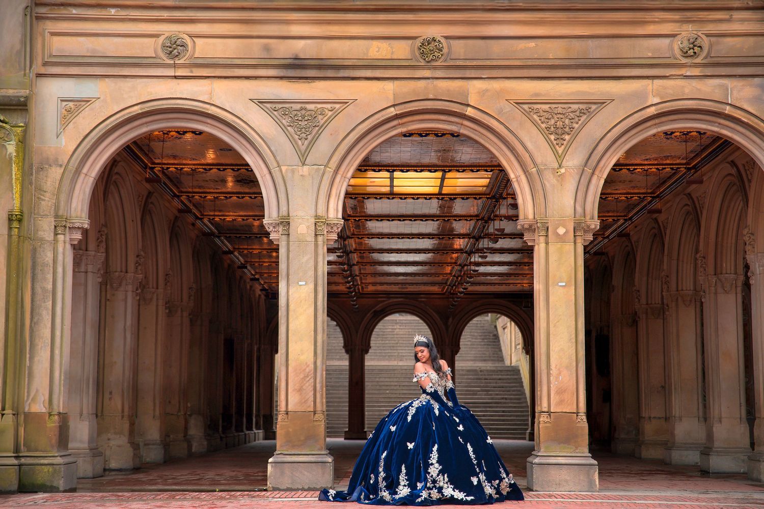 Elegant figure in a deep blue ballgown poses beneath ornate stone archways in Central Park's Bethesda Terrace arcade.