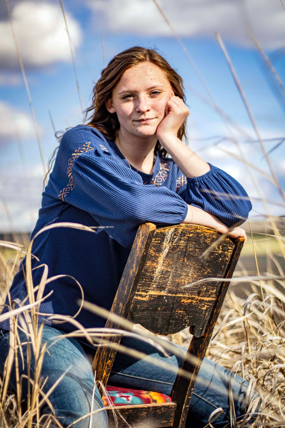 young woman sits backwards on rustic chair amid field of tall grass. cheek rests on her hand as she looks pleasantly toward the camera