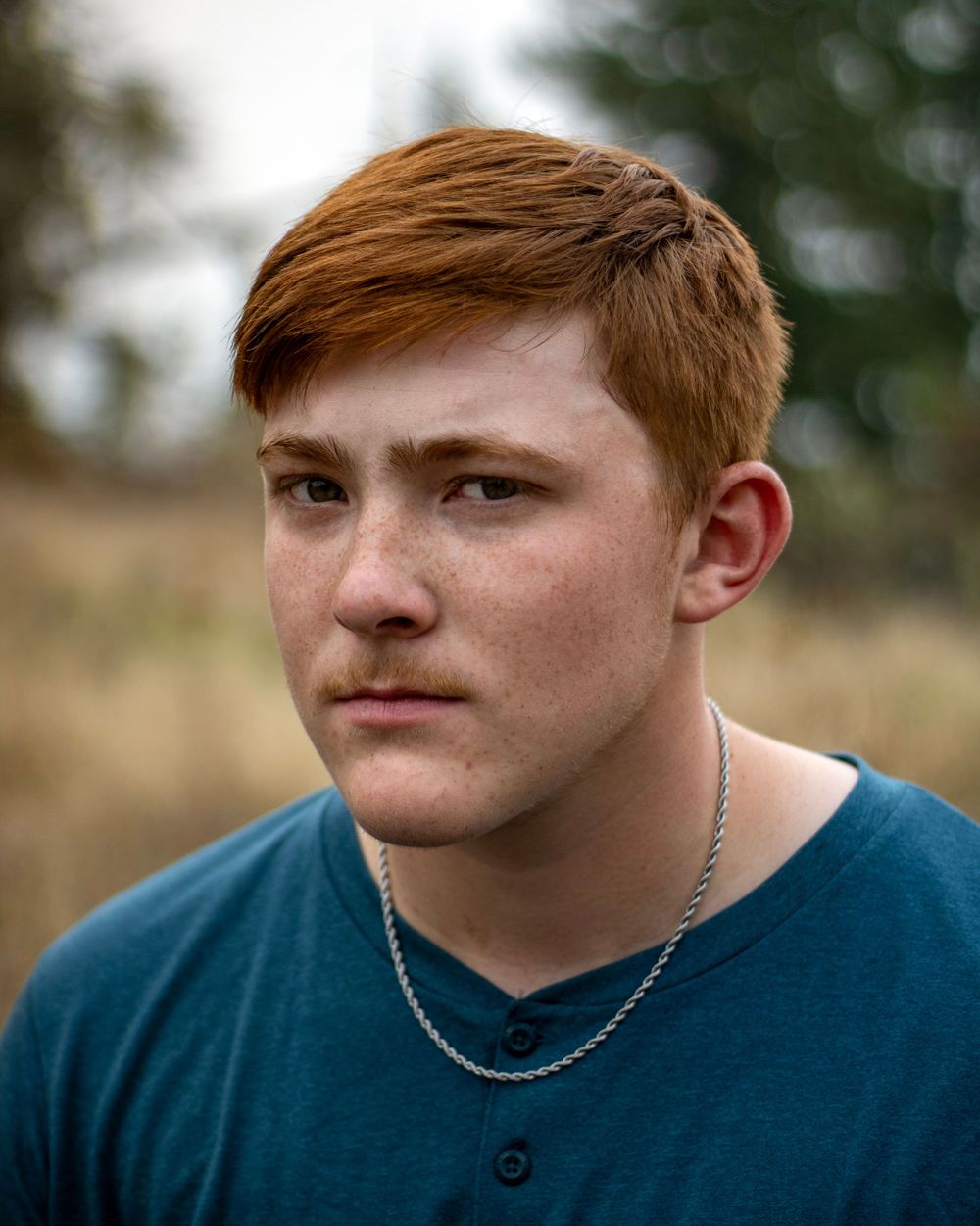 young man with red hair, teal shirt, and silver chain stares into camera. golden fields and green trees are behind him