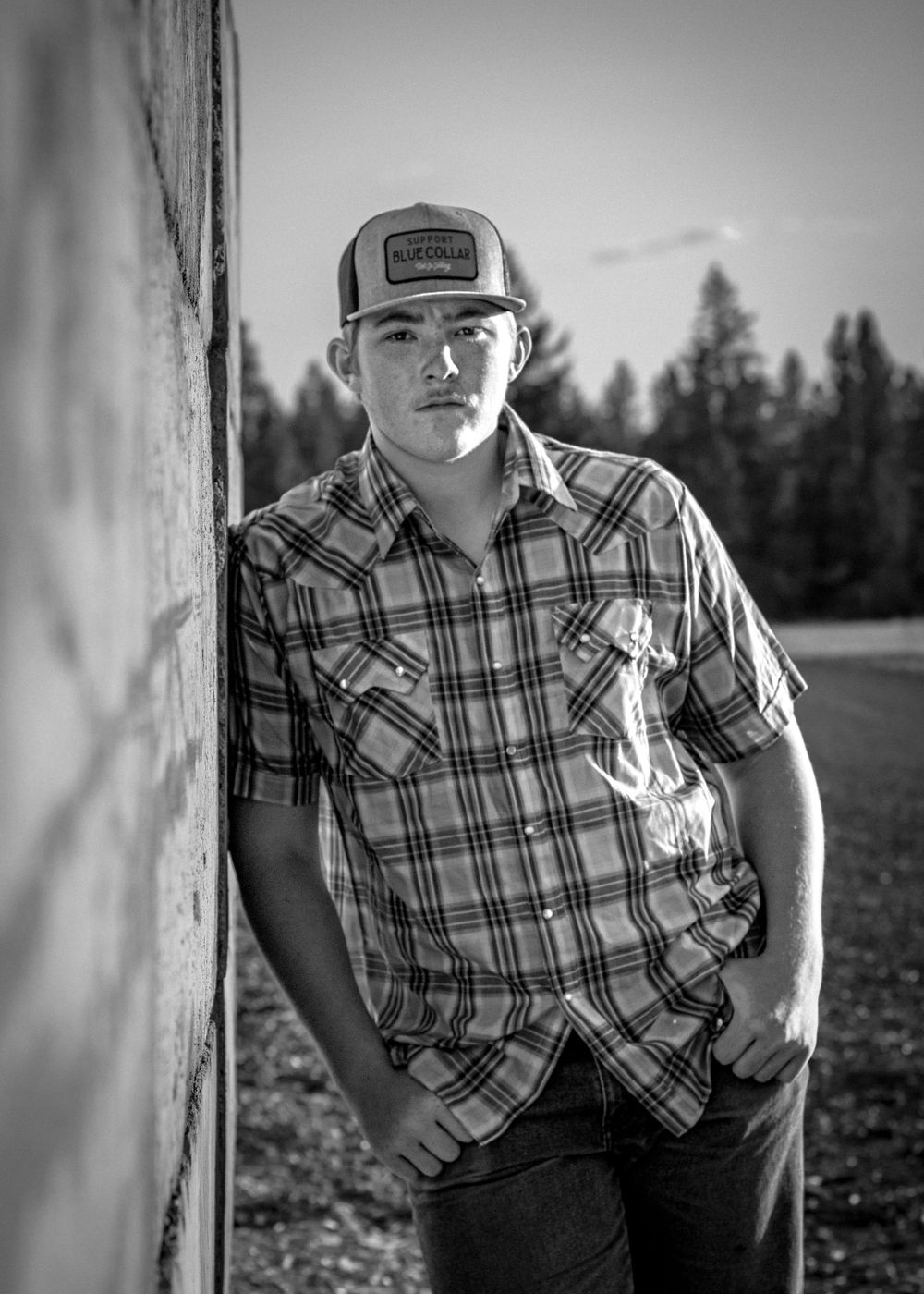 young man in plaid shirt leans shoulder against brick wall with hands in pockets, staring at the camera
