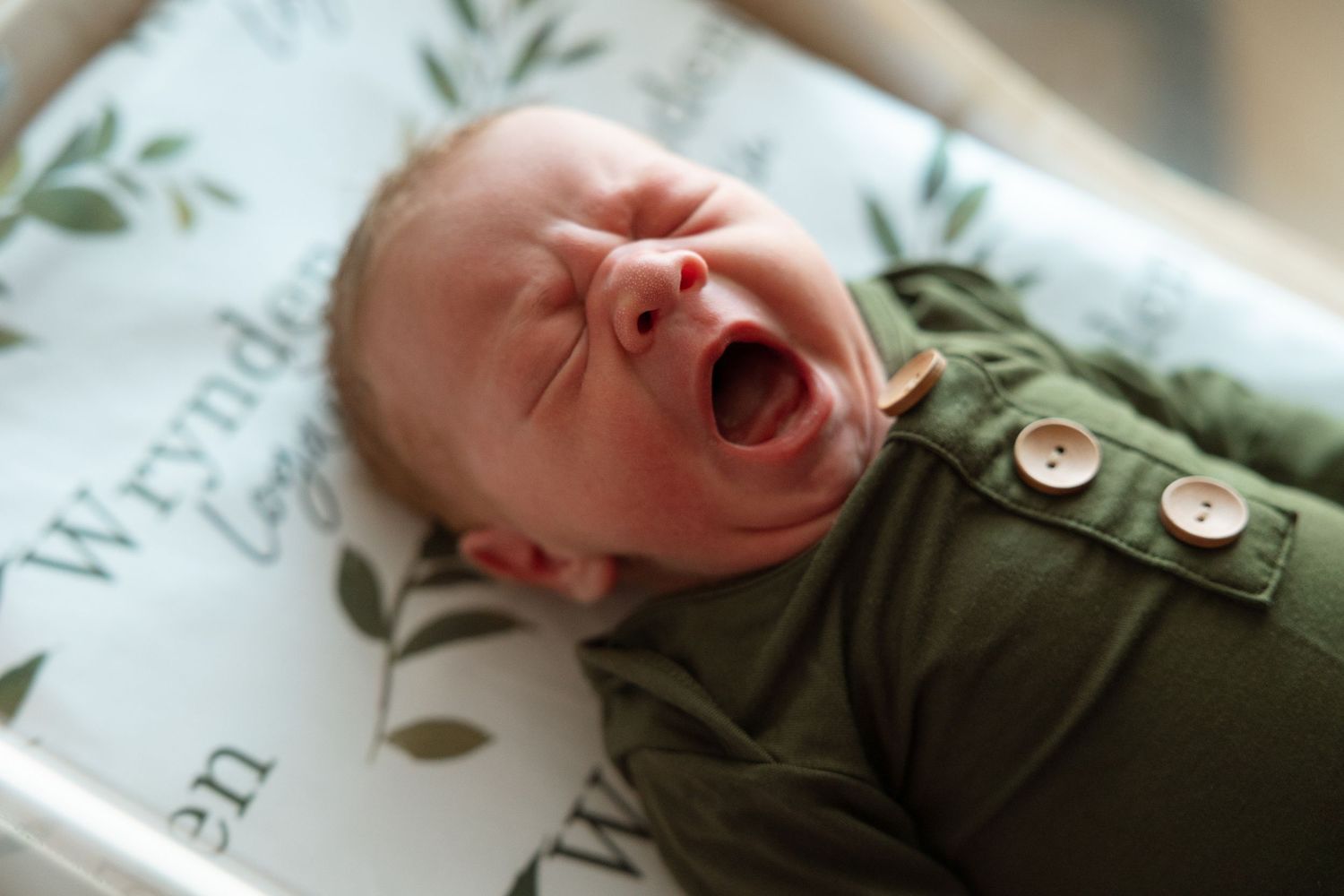 Newborn baby lying comfortably in cozy green onesie on white blanket, softly yawning.