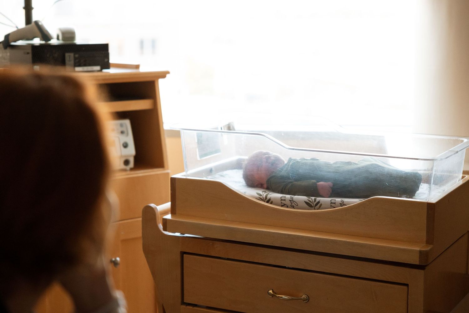 A clear plastic bassinet sits atop a wooden dresser in a nursery setting with soft natural lighting.