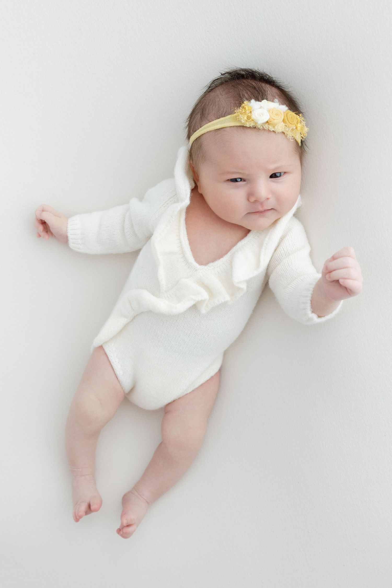 Baby wearing white bodysuit and yellow floral headband lies on white background.