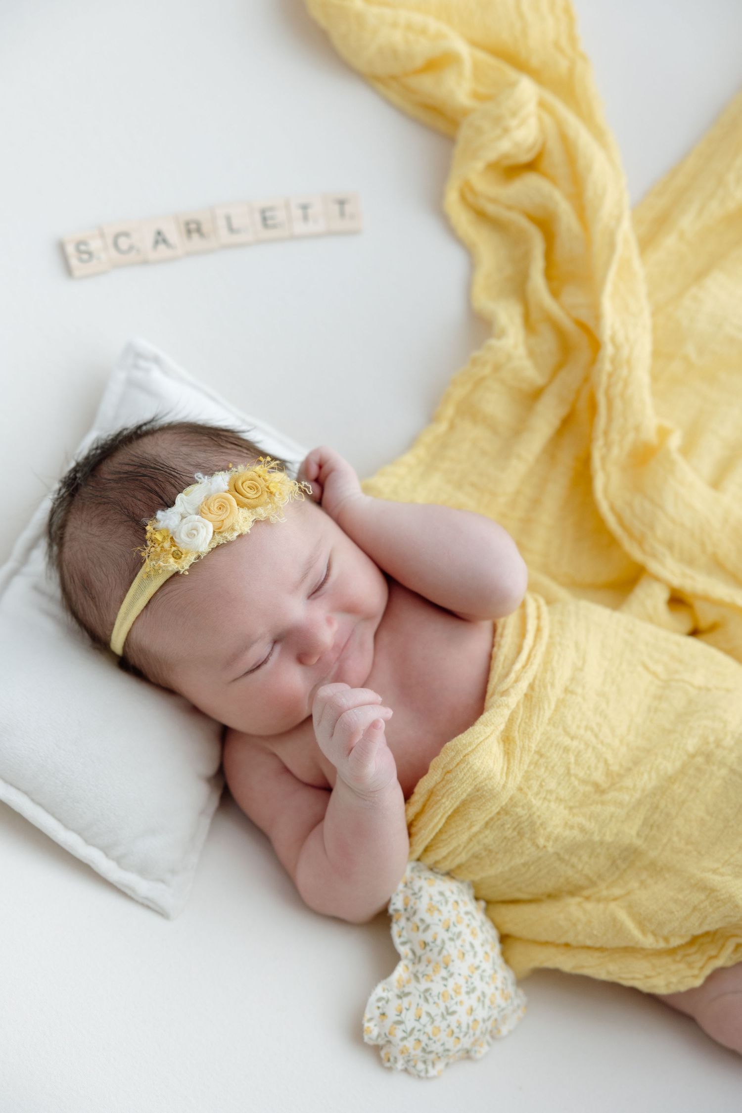 Sleeping newborn wrapped in soft yellow fabric wearing delicate floral headband during professional photo session.