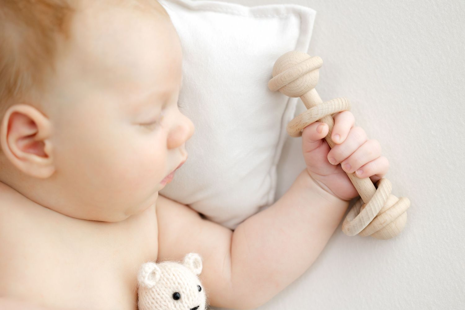 Sleeping baby holds wooden rattle while resting peacefully on white pillow with small teddy bear nearby.