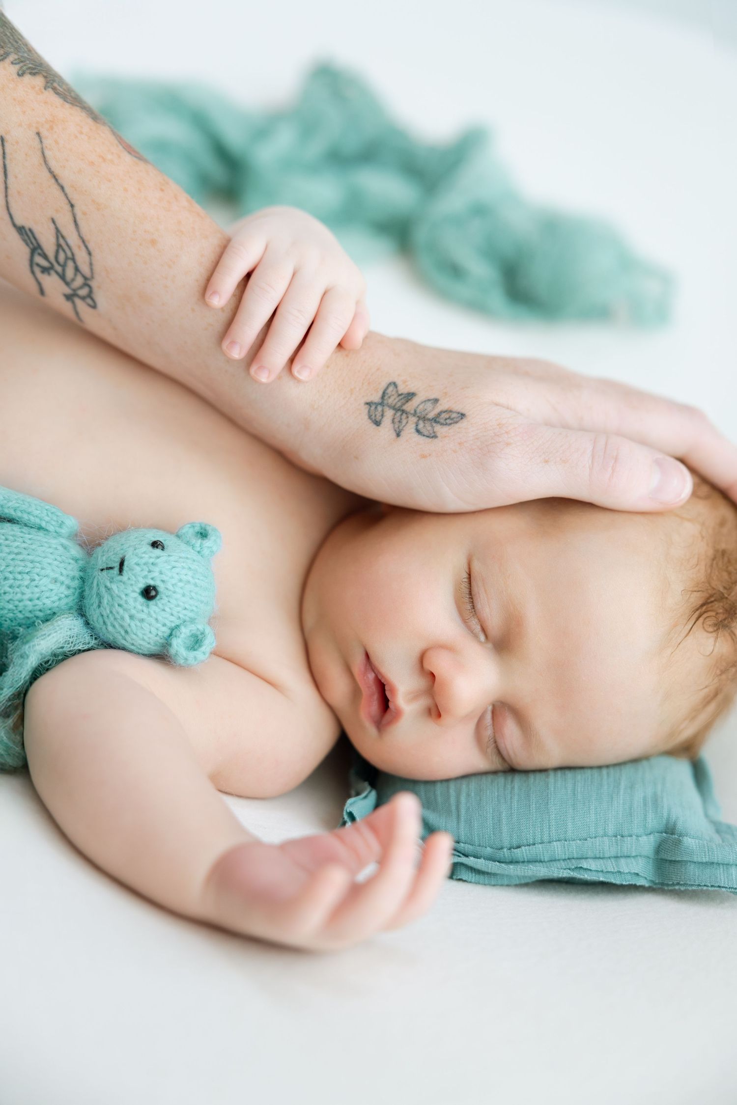 Sleeping infant with turquoise teddy bear rests peacefully next to tattooed arm on white surface.