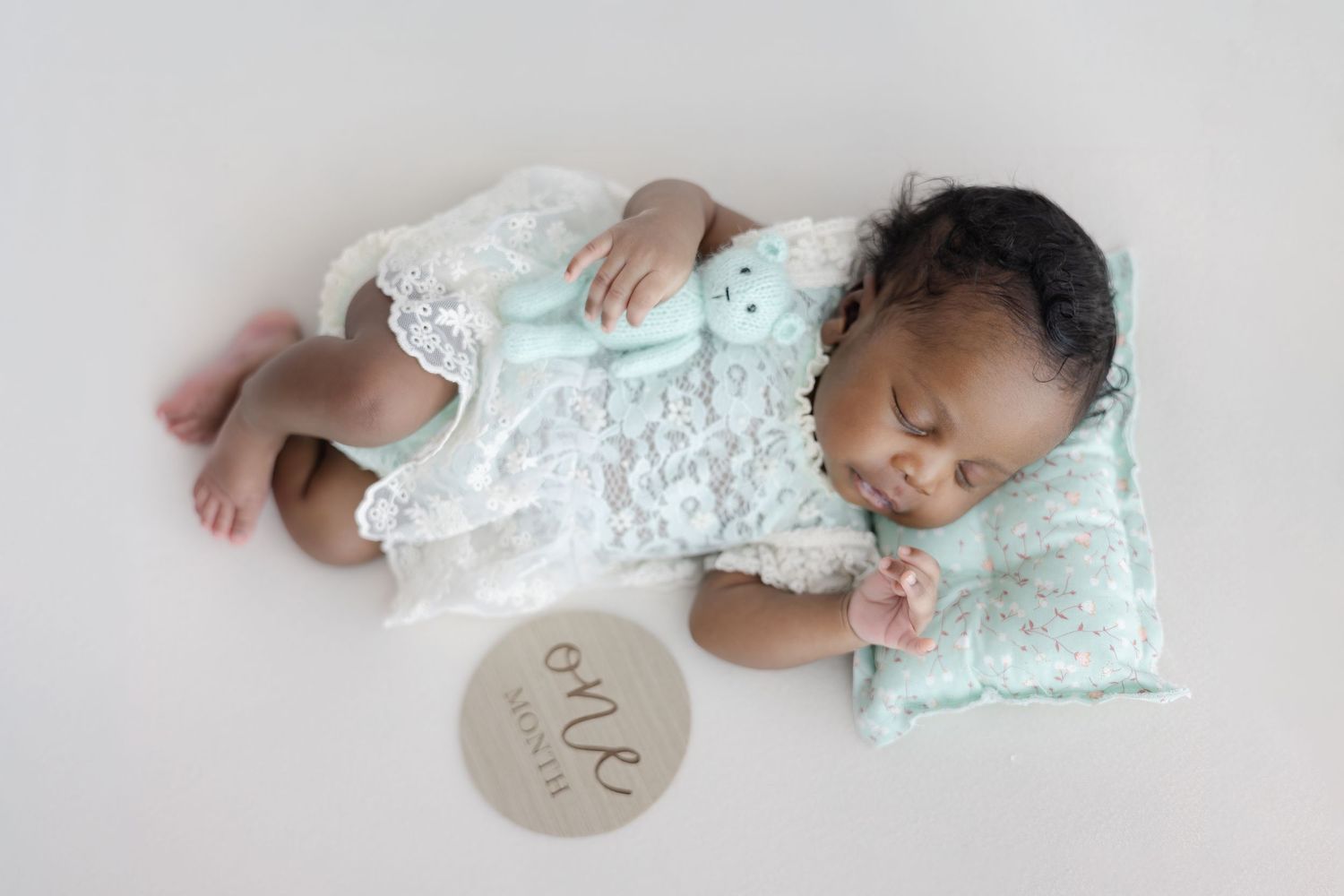 Sleeping baby wearing mint green outfit lies on white backdrop with wooden milestone marker in progressive photo series.