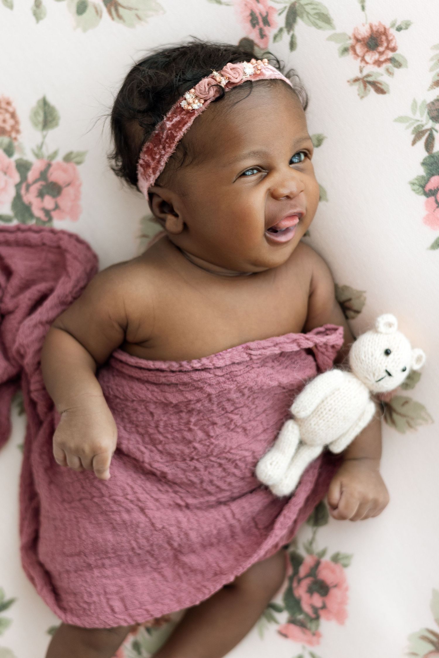 Sweet baby in pink dress smiles while holding white teddy bear against floral backdrop.
