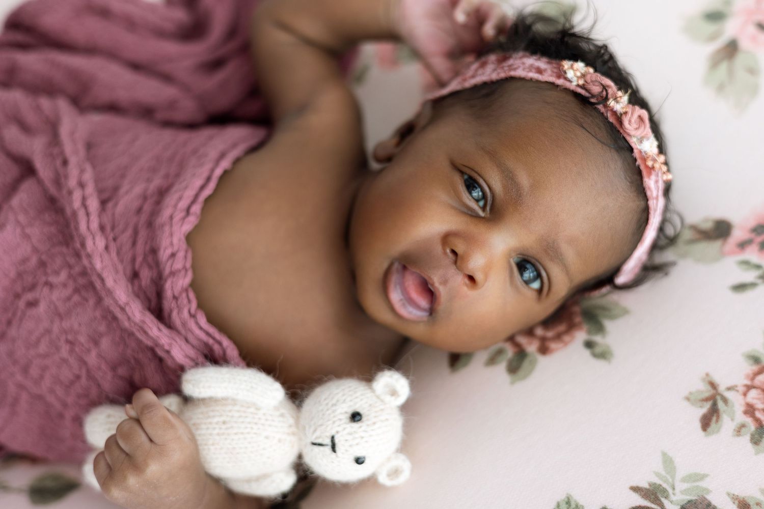 A newborn baby rests peacefully on pink bedding surrounded by delicate floral decorations and a small white teddy bear.