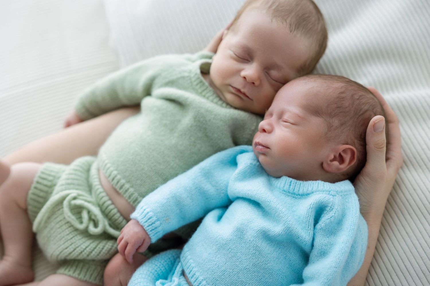 Sleeping newborn baby wrapped in soft mint green and blue knitted blankets lies peacefully on white bedding.