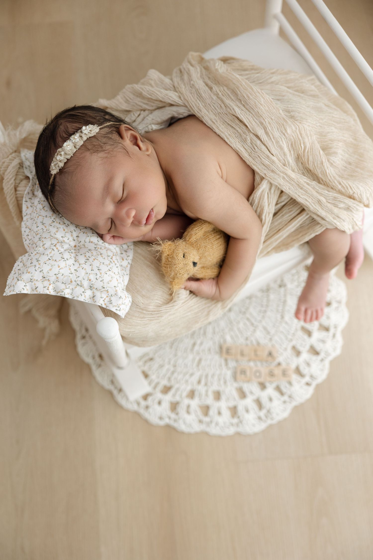 Newborn baby sleeping peacefully in a white basket with a teddy bear, floral pillow, and beige wrap.