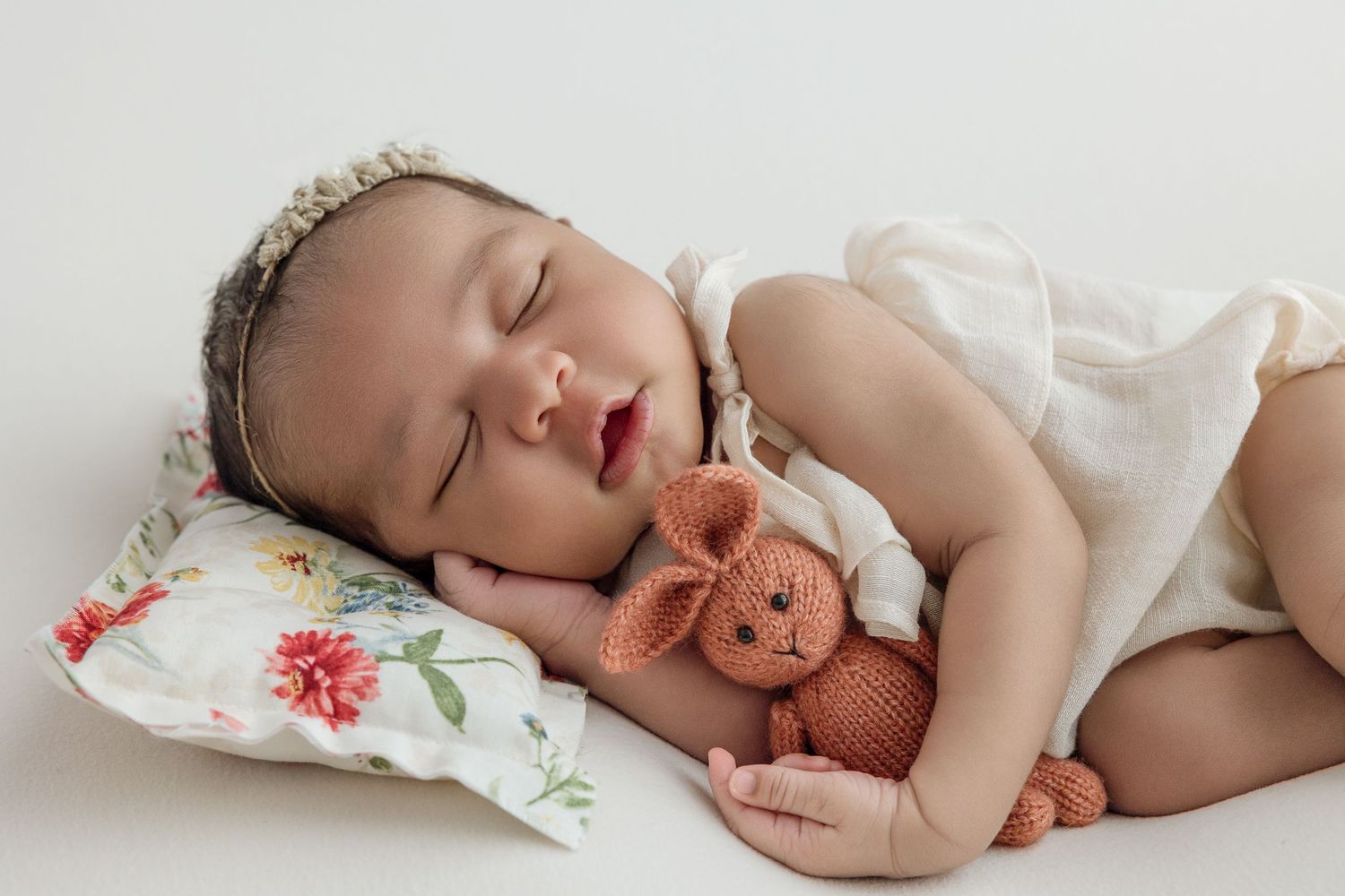 Sleeping newborn baby holding a knitted bunny toy, resting on a floral pillow in a white outfit.
