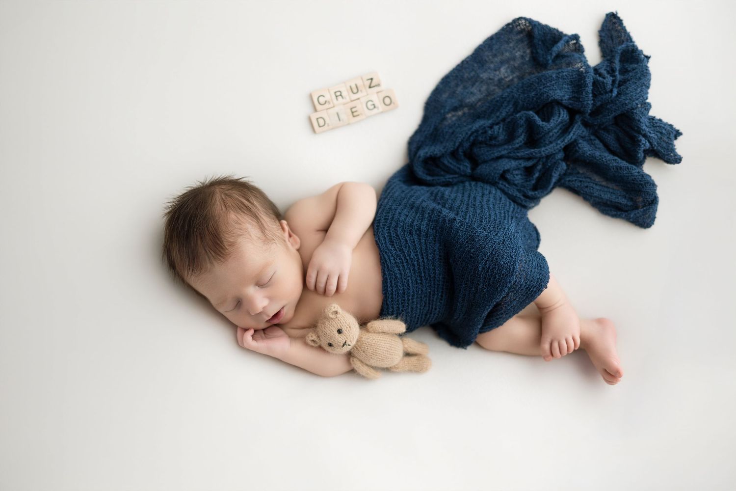 Newborn baby Cruz Diego sleeping peacefully wrapped in blue knit blanket with teddy bear on white background.
