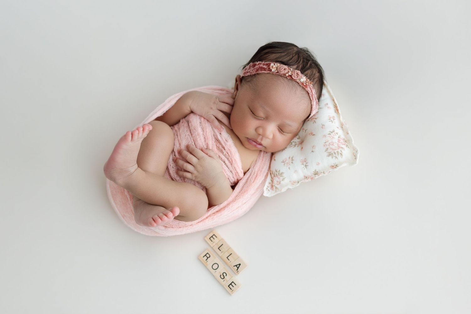 Newborn baby girl wrapped in pink swaddle sleeping on floral pillow with name tiles spelling Ella Rose.