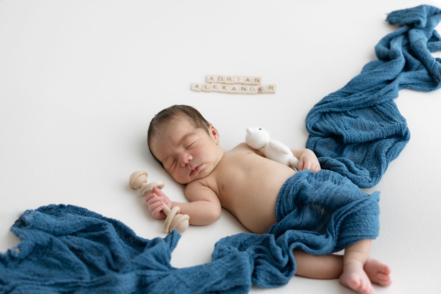 A newborn baby sleeps peacefully on white bedding while wrapped in a flowing blue blanket.