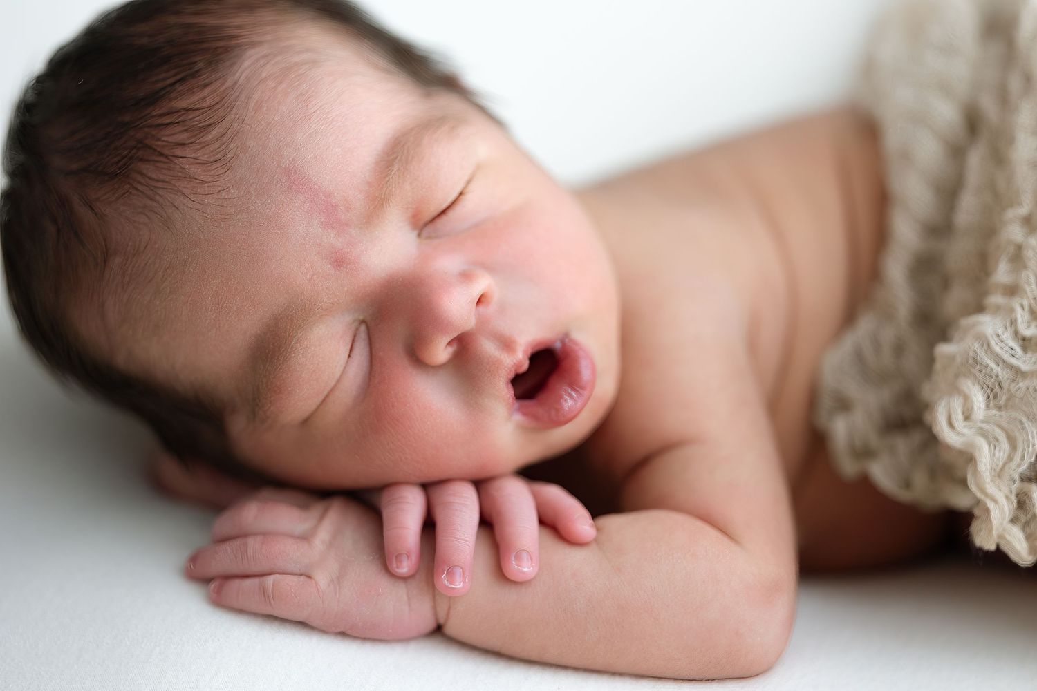 A sleeping newborn baby wrapped in soft beige lace fabric lies peacefully during a professional photo session.